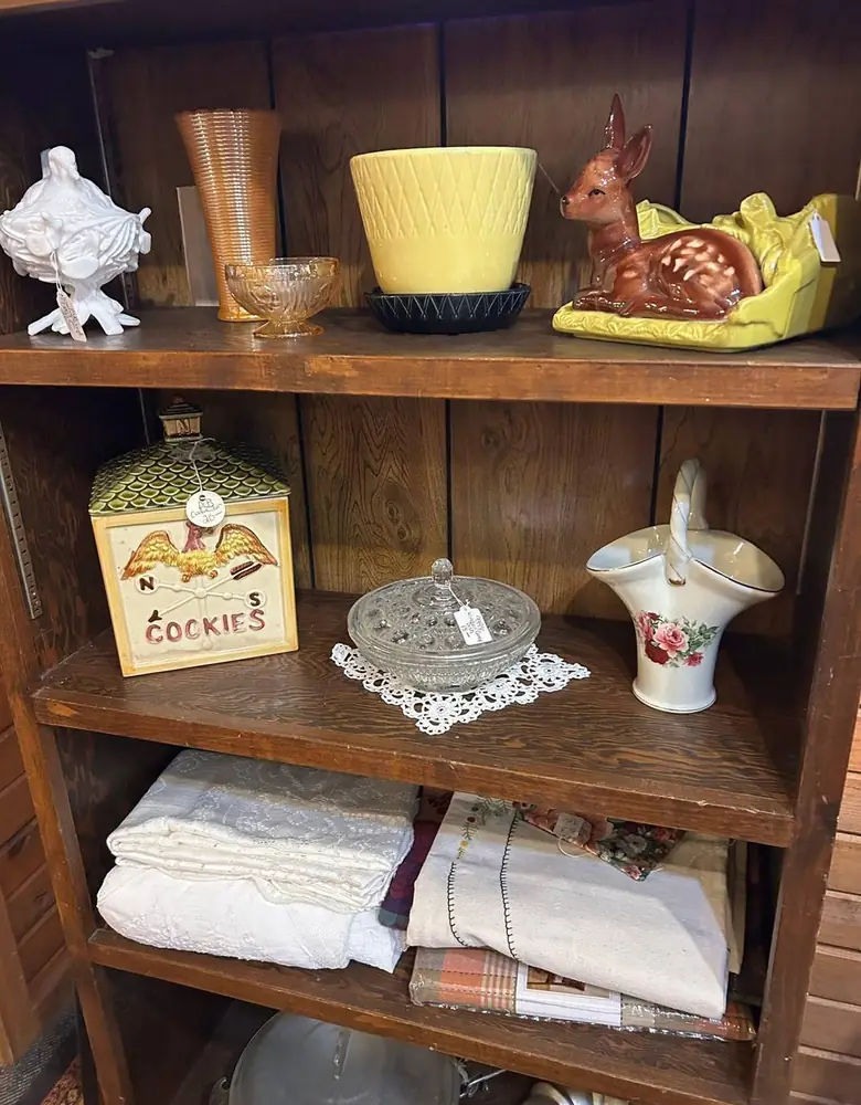 Wooden shelf at Chaintiques with a vintage cookie tin, a pressed glass dish, a flowered porcelain basket, and folded linens